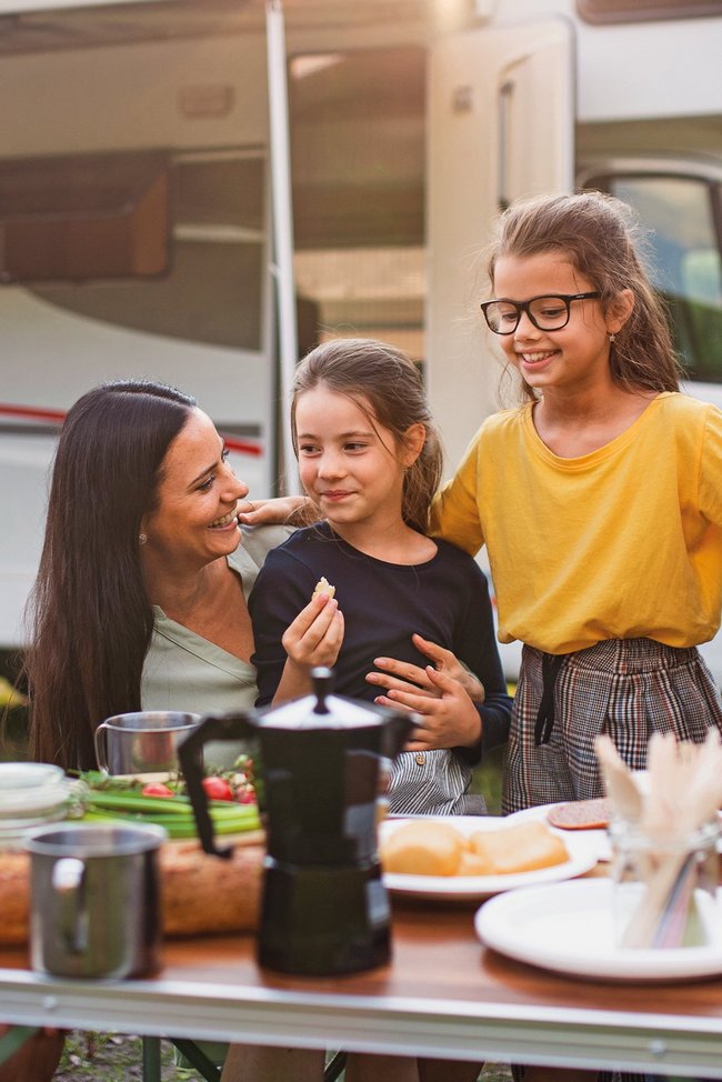 Familie isst am Tisch im Freien, mit einem Wohnwagen im Hintergrund.
